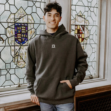 College-aged man wearing the Pi Kappa Phi embroidered black hoodie, standing in front of stained glass windows with fraternity-style crest designs.