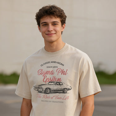 Young man wearing a vintage-style beige graphic T-shirt featuring a retro muscle car illustration and cursive text, standing outdoors with a neutral blurred background.
