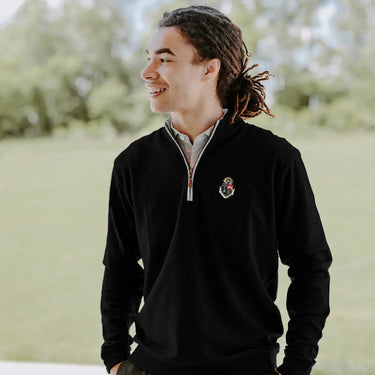 College-aged man smiles and looks off to the side while wearing a black quarter-zip pullover embroidered with a fraternity crest. He stands outdoors in a bright, grassy setting.