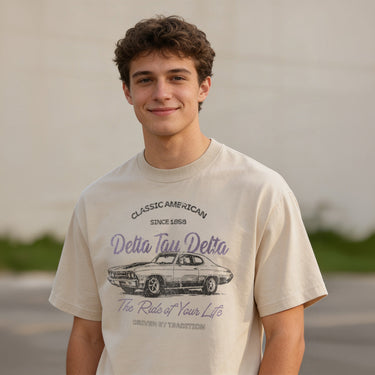Young man wearing a vintage-style beige graphic T-shirt featuring a retro muscle car illustration and cursive text, standing outdoors with a neutral blurred background.