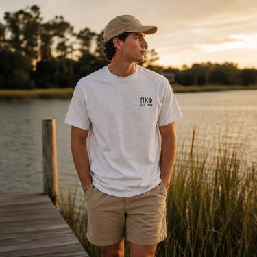 Young man wearing a white short-sleeve T-shirt with a small left chest graphic while standing on a lakeside dock at sunset.
