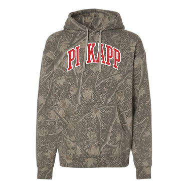 College-aged male wearing the fraternity Deadwood Camo Hoodie with red and white Pi Kappa Phi lettering, seated outdoors in a wooded setting.