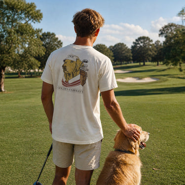 Lifestyle photo of a college student wearing a golf-themed fraternity t-shirt on a golf course with a golden retriever. Ivory Comfort Colors tee features a “Golden Fairways” back graphic with a golden retriever in golf apparel and golf clubs, perfect for fraternity golf tournaments, campus events, and casual Greek life apparel.
