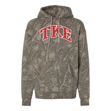 College-aged male wearing the fraternity Deadwood Camo Hoodie with red and white Tau Kappa Epsilon lettering, seated outdoors in a wooded setting.