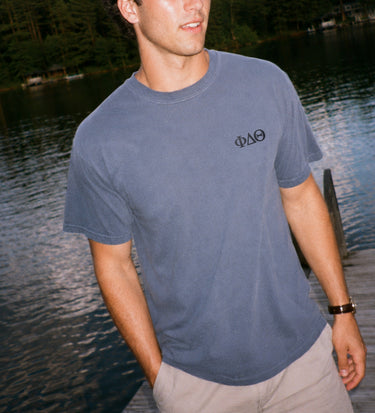 Young man wearing a blue Comfort Colors style t-shirt by the lake, showing the front chest design