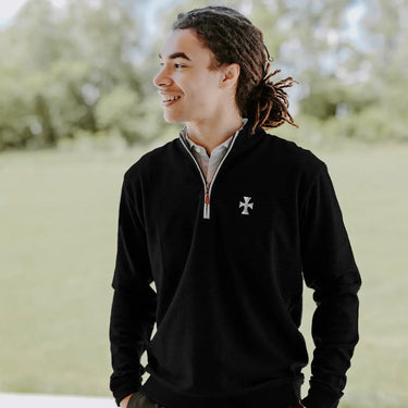 College-aged man smiles and looks off to the side while wearing a black quarter-zip pullover embroidered with a fraternity crest. He stands outdoors in a bright, grassy setting.