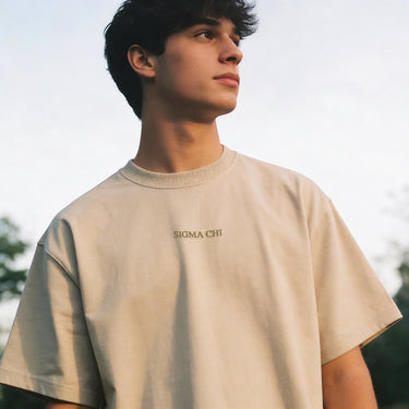 Man wearing a beige crewneck t-shirt with subtle embroidered text on the chest, photographed outdoors.