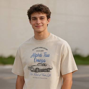 Young man wearing a vintage-style beige graphic T-shirt featuring a retro muscle car illustration and cursive text, standing outdoors with a neutral blurred background.