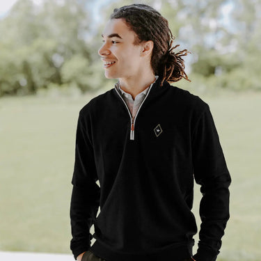 College-aged man smiles and looks off to the side while wearing a black quarter-zip pullover embroidered with a fraternity crest. He stands outdoors in a bright, grassy setting.