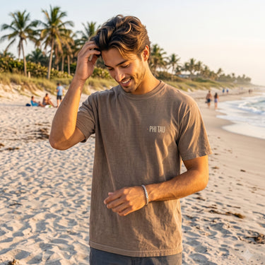 Model wearing the brown Surf Club Social T-shirt with a small front chest logo on a beach.