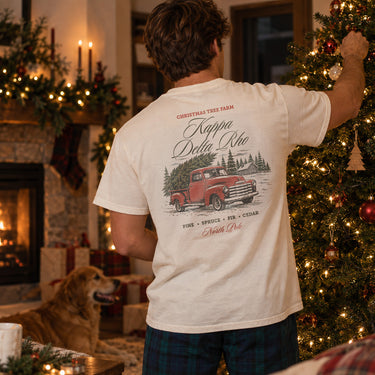 College-aged man wearing a limited edition Tree Farm Holiday Tee in a cozy Christmas setting with a decorated tree and fireplace.
