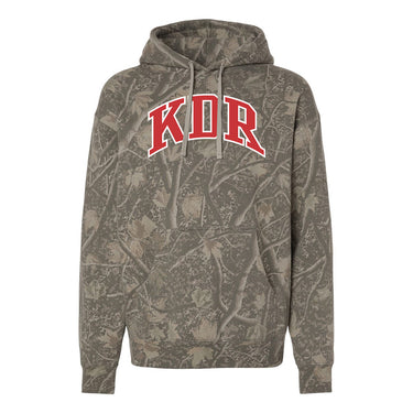 College-aged male wearing the fraternity Deadwood Camo Hoodie with red and white Kappa Delta Rho lettering, seated outdoors in a wooded setting.