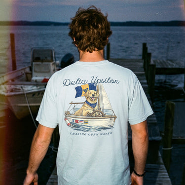 Young man standing on a dock at sunset wearing a light blue t-shirt featuring a golden retriever steering a sailboat with a flag and the phrase “Chasing Open Water” on the back.