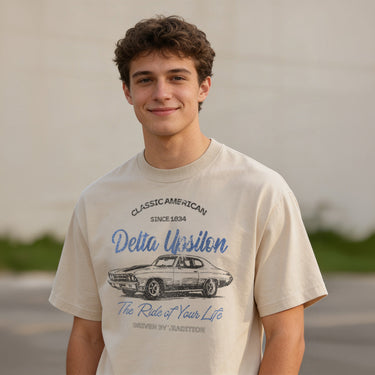 Young man wearing a vintage-style beige graphic T-shirt featuring a retro muscle car illustration and cursive text, standing outdoors with a neutral blurred background.