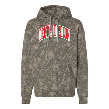 College-aged male wearing the fraternity Deadwood Camo Hoodie with red and white Delta Uspilon lettering, seated outdoors in a wooded setting.