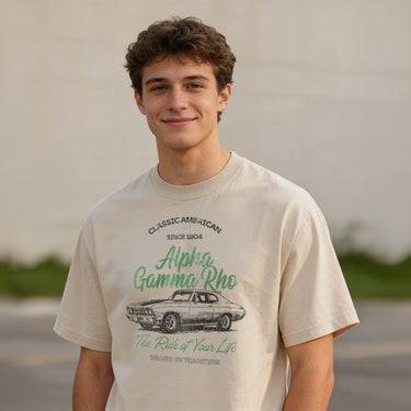 Young man wearing a vintage-style beige graphic T-shirt featuring a retro muscle car illustration and cursive text, standing outdoors with a neutral blurred background.