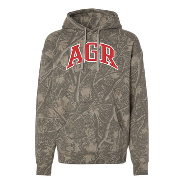 College-aged male wearing the fraternity Deadwood Camo Hoodie with red and white Alpha Gamma Rho lettering, seated outdoors in a wooded setting.